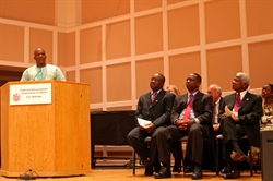 [ai] A speaker at a podium delivers a speech, while four seated men listen attentively. The setting appears to be a formal event, with a wooden stage and a banner reading 'Truth and Reconciliation Commission of Liberia'.